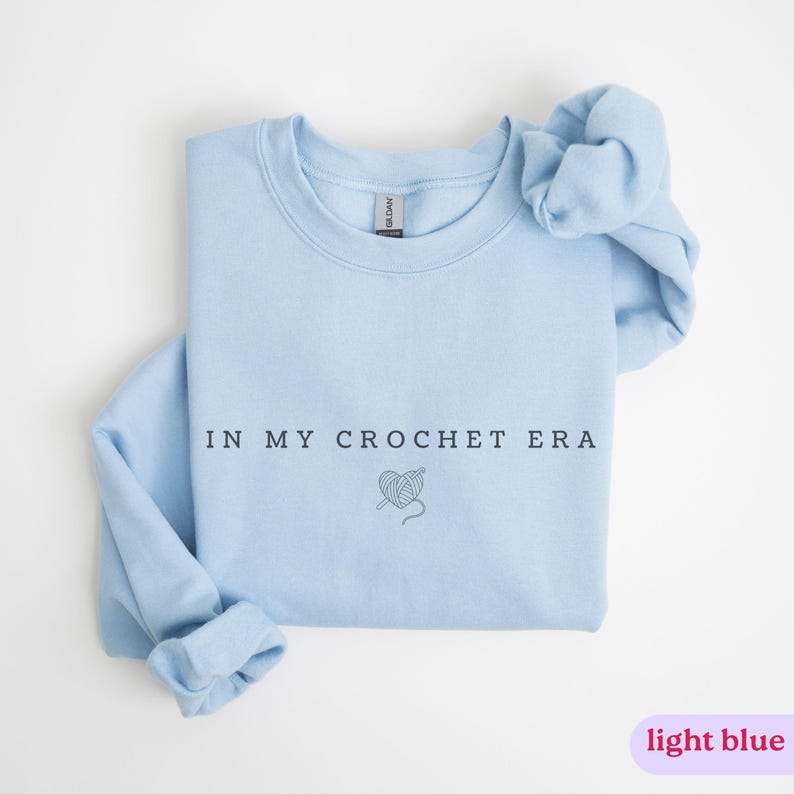 Puede incluir: Sudadera azul claro con el texto "In My Crochet Era" y un peque&ntilde;o gr&aacute;fico de un coraz&oacute;n con hilo y un ganchillo.