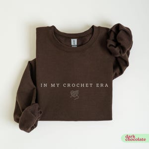 Puede incluir: Una sudadera de color marr&oacute;n chocolate oscuro con el texto "In My Crochet Era" y un peque&ntilde;o gr&aacute;fico de un coraz&oacute;n con hilo y un ganchillo.