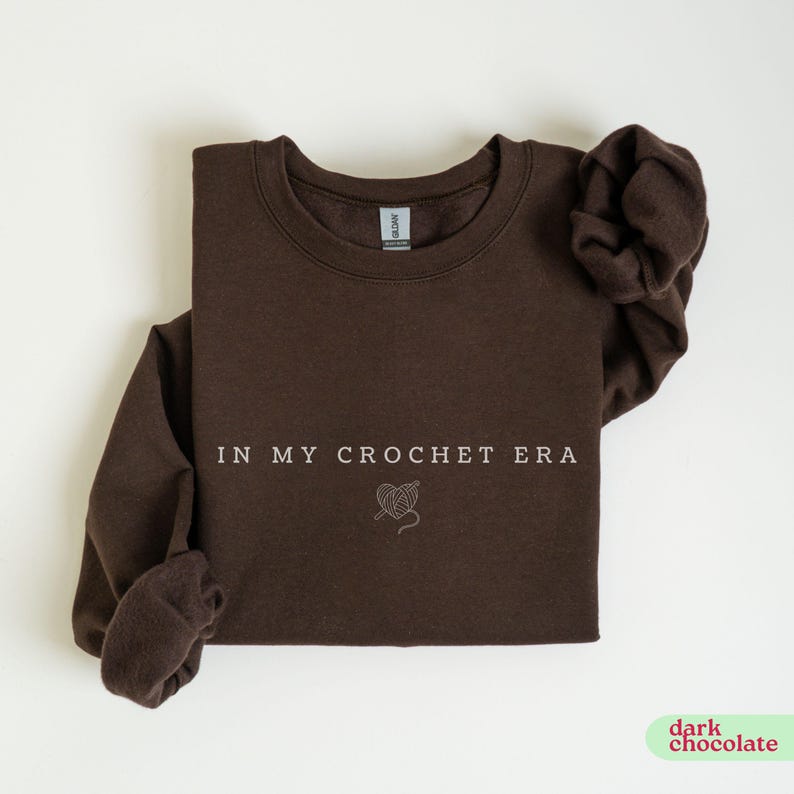 Puede incluir: Una sudadera de color marr&oacute;n chocolate oscuro con el texto "In My Crochet Era" y un peque&ntilde;o gr&aacute;fico de un coraz&oacute;n con hilo y un ganchillo.