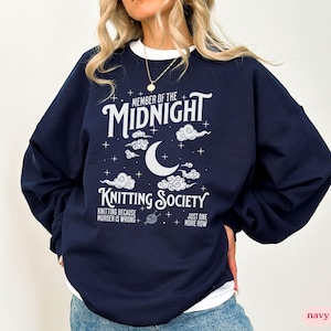 Puede incluir: Sudadera azul marino con texto blanco que dice "MEMBER OF THE MIDNIGHT KNITTING SOCIETY". El diseño incluye una luna creciente, nubes y estrellas. También se incluye el texto "KNITTING BECAUSE MURDER IS WRONG" y "JUST ONE MORE ROW".