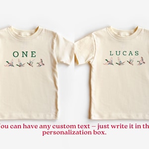 Peut inclure: Deux t-shirts cr&egrave;me pour tout-petits. L'un porte le mot "ONE" en vert au-dessus d'une rang&eacute;e de canards en vol. L'autre porte le nom "LUCAS" en vert au-dessus d'une rang&eacute;e de canards en vol. Le texte "You can have any custom text" est en bas.