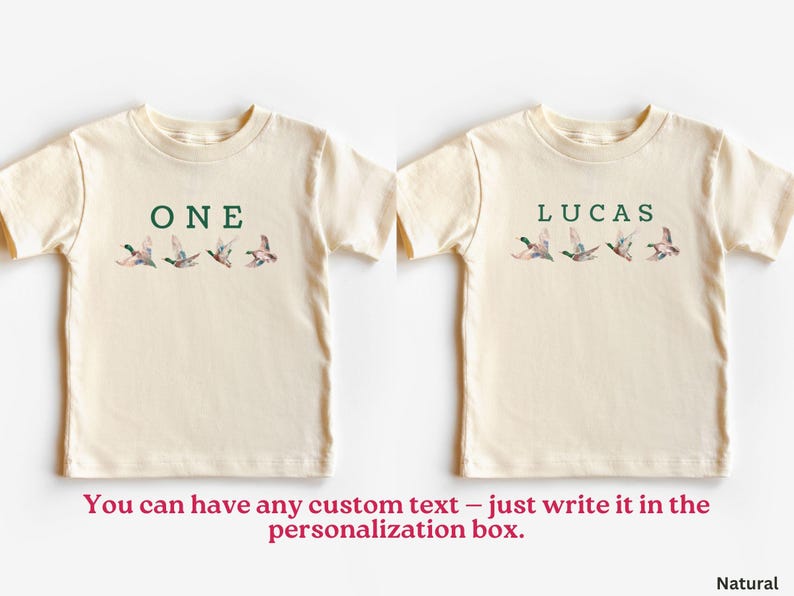 Peut inclure: Deux t-shirts cr&egrave;me pour tout-petits. L'un porte le mot "ONE" en vert au-dessus d'une rang&eacute;e de canards en vol. L'autre porte le nom "LUCAS" en vert au-dessus d'une rang&eacute;e de canards en vol. Le texte "You can have any custom text" est en bas.