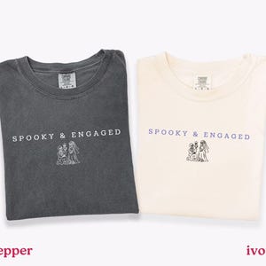 May include: Two crew neck t-shirts in pepper gray and ivory. Each shirt features the text SPOOKY & ENGAGED in a simple font above a line art illustration of a skeleton proposing to a skeleton bride. The shirts have a classic fit and short sleeves.