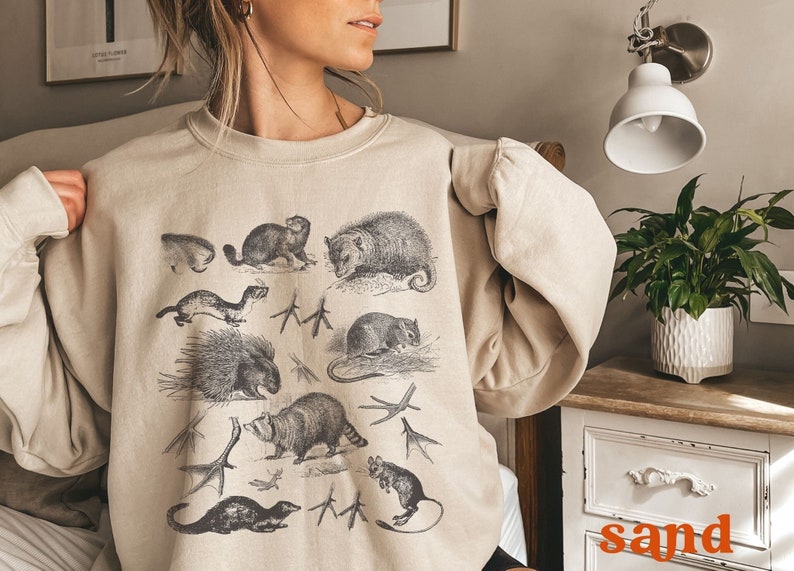 May include: A beige sweatshirt featuring a vintage illustration of various animals, including a raccoon, a porcupine, a weasel, and an opossum. The animals are printed in black and white on a light beige background.