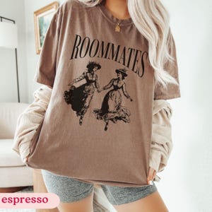 May include: A brown t-shirt with the text "ROOMMATES" printed in black. The shirt features a graphic of two women in vintage dresses holding hands.