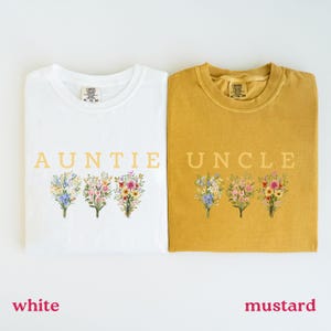 May include: White and mustard-colored t-shirts with the words "AUNTIE" and "UNCLE" printed in gold. Each shirt features a floral bouquet design in pastel colors. The shirts are a Comfort Colors brand.
