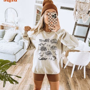 May include: A beige sweatshirt featuring a graphic print of various animals, including a raccoon, a porcupine, a weasel, and a mouse. The sweatshirt has a relaxed fit and a crew neckline.