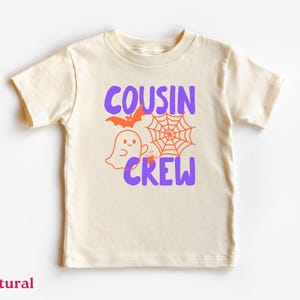May include: A natural-colored short-sleeve t-shirt featuring a graphic print with the purple text COUSIN CREW. The design includes an orange bat, a spider web, a small ghost, and a spider, all centered on the front of the garment.