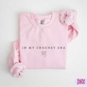 Puede incluir: Una sudadera rosa claro con el texto "In My Crochet Era" y un peque&ntilde;o gr&aacute;fico de un coraz&oacute;n con hilo y un gancho de ganchillo.