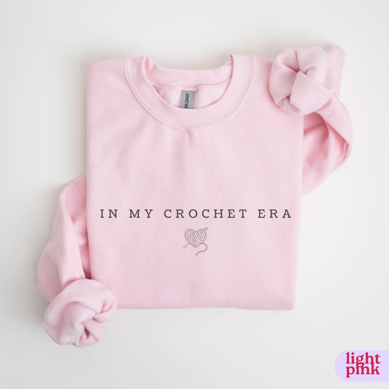 Puede incluir: Una sudadera rosa claro con el texto "In My Crochet Era" y un peque&ntilde;o gr&aacute;fico de un coraz&oacute;n con hilo y un gancho de ganchillo.