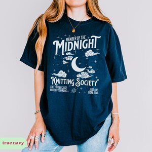 May include: A navy blue t-shirt with white text and graphics. The shirt reads "MEMBER OF THE MIDNIGHT KNITTING SOCIETY" with a moon and cloud design. Additional text includes "KNITTING BECAUSE MURDER IS WRONG" and "JUST ONE MORE ROW."