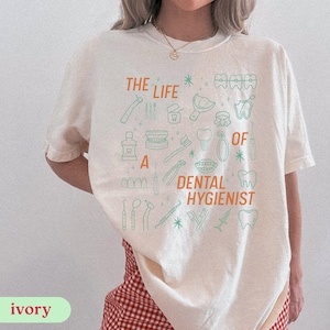 May include: Ivory-colored t-shirt with the text "THE LIFE OF A DENTAL HYGIENIST" in orange and green. The shirt features dental-themed illustrations, including teeth, toothbrushes, and dental tools. The shirt is made of a soft material.
