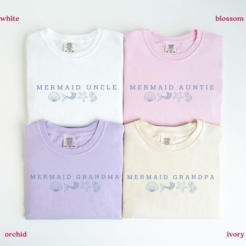 Puede incluir: Cuatro camisetas dobladas en blanco, rosa flor, morado orqu&iacute;dea y marfil. Cada camiseta tiene las palabras "Mermaid Uncle", "Mermaid Auntie", "Mermaid Grandma" y "Mermaid Grandpa" impresas en azul, junto con gr&aacute;ficos de conchas marinas, cola de sirena, estrella de mar e hipocampo.