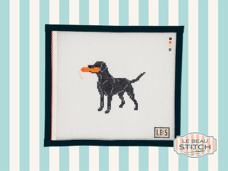 Puede incluir: Lienzo de punto de aguja con un Labrador Retriever negro sosteniendo un juguete naranja. El perro est&aacute; cosido sobre lienzo blanco, enmarcado por un borde verde oscuro con acentos naranjas. El dise&ntilde;o incluye el texto "LBS" y "LE BEAU STITCH".