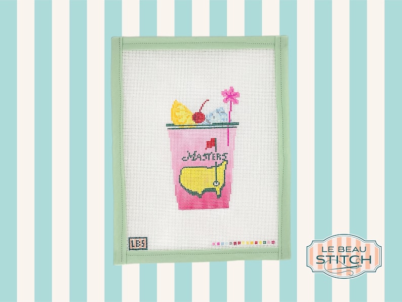 May include: A needlepoint canvas with a green border featuring a pink cocktail glass with a lemon wedge, cherry, and a pink flower. The glass has the words "Masters" and a yellow golf course with a flag.