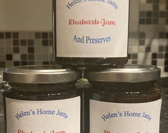 May include: Three jars of rhubarb jam with silver lids are stacked. Each jar features a white label with the text "Helen's Home Jams," "Rhubarb Jam," and "And Preserves" in blue and red fonts. The jars are filled with a dark preserve.