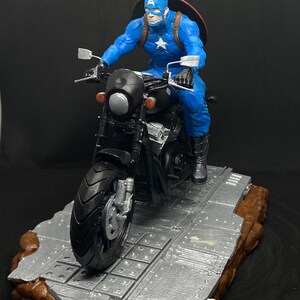 May include: Captain America action figure riding a black motorcycle on a gray and brown base. The figure is wearing a blue and red costume with a red, white, and blue star on the chest. The motorcycle has a black seat and handlebars. The base is textured and has a gray and brown color scheme. The word "Pigeon" is written in white on the base.