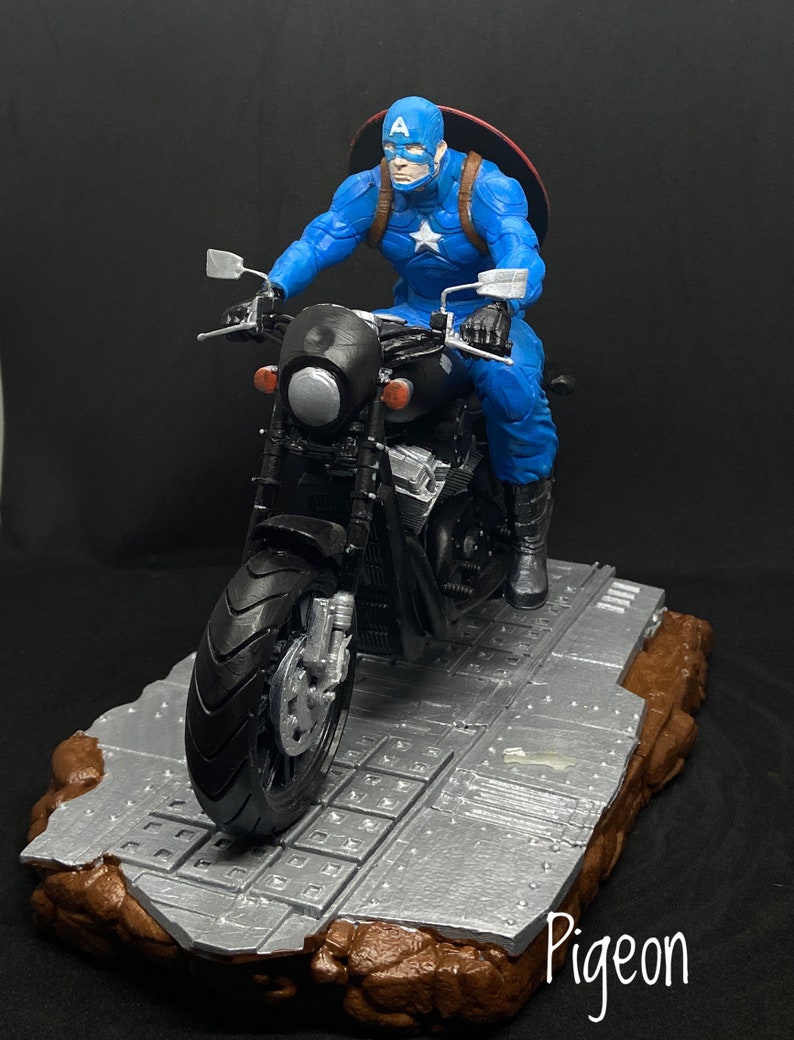 May include: Captain America action figure riding a black motorcycle on a gray and brown base. The figure is wearing a blue and red costume with a red, white, and blue star on the chest. The motorcycle has a black seat and handlebars. The base is textured and has a gray and brown color scheme. The word "Pigeon" is written in white on the base.
