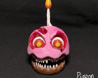 May include: A pink and brown animatronic head with a single lit candle on top. The animatronic has red eyes with yellow centres and a toothy grin.