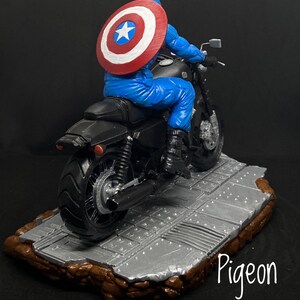 May include: A black motorcycle with a figure wearing a blue and red costume with a star on the chest, riding the motorcycle. The motorcycle is on a gray platform with a brown base. The text "Pigeon" is written on the base.