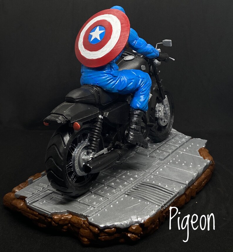 May include: A black motorcycle with a figure wearing a blue and red costume with a star on the chest, riding the motorcycle. The motorcycle is on a gray platform with a brown base. The text "Pigeon" is written on the base.