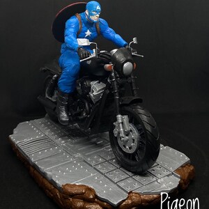 May include: A blue and red Captain America action figure riding a black motorcycle on a gray and brown base. The figure is holding the motorcycle handlebars and wearing a helmet. The motorcycle has a black seat and a black exhaust pipe.