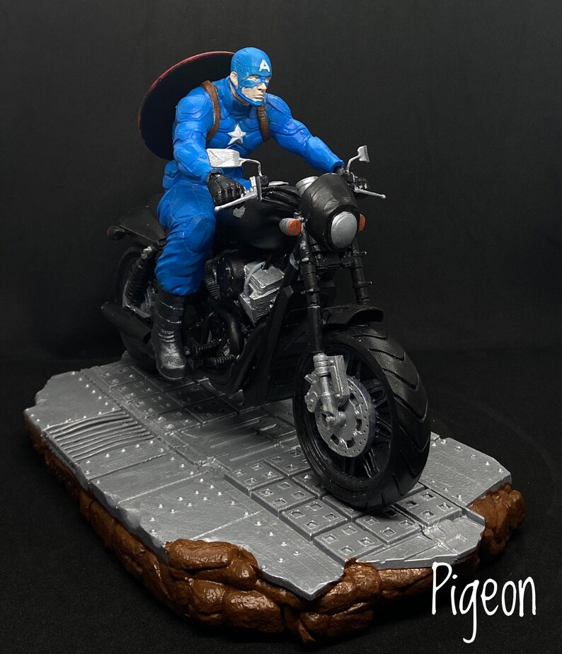 May include: A blue and red Captain America action figure riding a black motorcycle on a gray and brown base. The figure is holding the motorcycle handlebars and wearing a helmet. The motorcycle has a black seat and a black exhaust pipe.