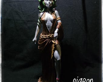 Puede incluir: Estatua de Medusa con piel plateada, cabello de serpiente verde y corona dorada. Viste un vestido color bronce y sostiene una daga dorada. La estatua tiene un diseño detallado y se alza sobre una superficie oscura.