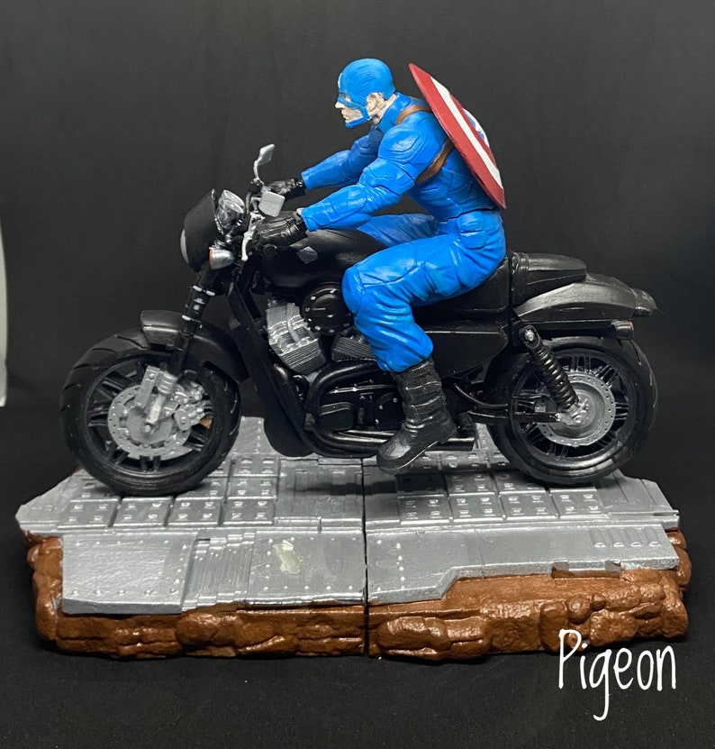 May include: A black motorcycle with a red, white, and blue shield on the back. A figure in a blue and red costume is riding the motorcycle. The motorcycle is on a gray and brown base.