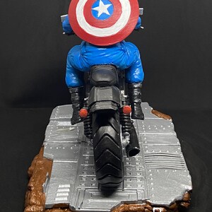 May include: A blue and red Captain America action figure riding a black motorcycle with a silver and brown base. The figure is wearing a red, white, and blue helmet and has a red, white, and blue shield on his back.