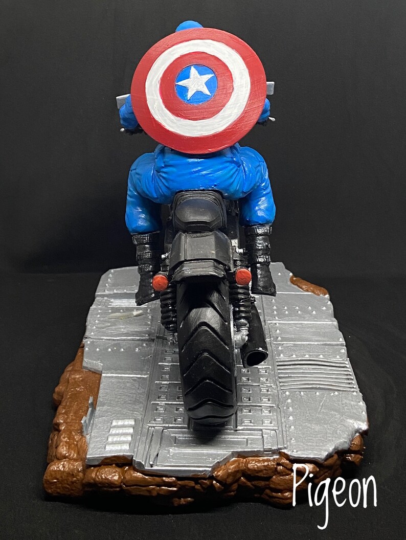 May include: A blue and red Captain America action figure riding a black motorcycle with a silver and brown base. The figure is wearing a red, white, and blue helmet and has a red, white, and blue shield on his back.