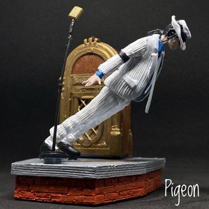 May include: A white figurine of a singer wearing a white suit and a white hat, leaning forward with a microphone stand behind them. The figurine is on a stage with a brick base and a gold jukebox behind them.