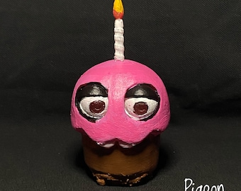 May include: A pink cupcake character with black eyes, a white candle with a yellow flame, and a brown cupcake base. The character is a handmade figurine.