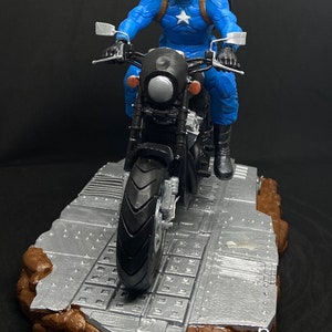 May include: A blue and red Captain America action figure riding a black motorcycle on a gray and brown platform. The figure is wearing a helmet and holding the handlebars. The motorcycle has a black seat and a silver headlight. The platform is made of a textured material and has a brown base.