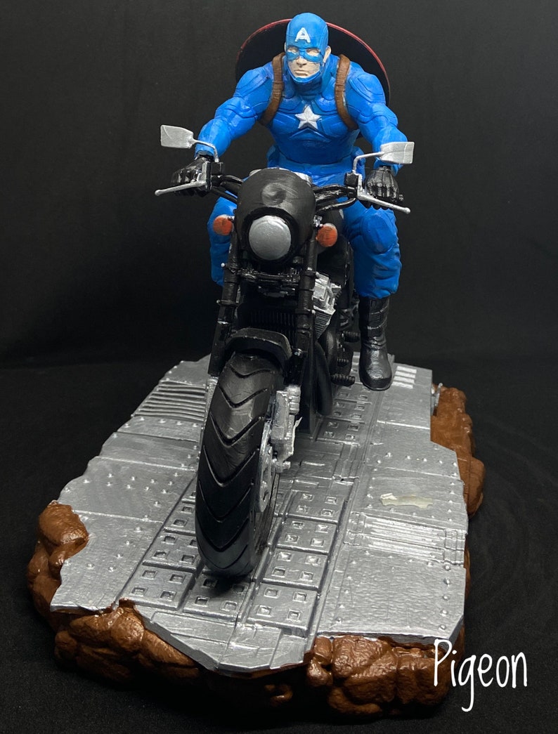 May include: A blue and red Captain America action figure riding a black motorcycle on a gray and brown platform. The figure is wearing a helmet and holding the handlebars. The motorcycle has a black seat and a silver headlight. The platform is made of a textured material and has a brown base.