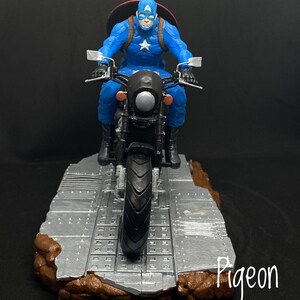 May include: Captain America action figure riding a black motorcycle with a silver base. The figure is wearing a blue and red costume with a white star on the chest. The motorcycle has a silver headlight and a silver base.