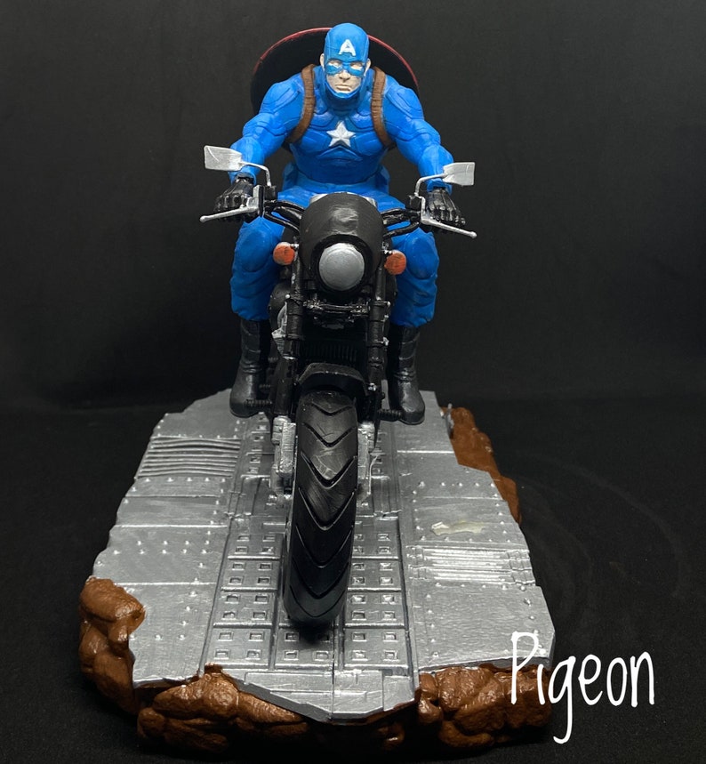 May include: Captain America action figure riding a black motorcycle with a silver base. The figure is wearing a blue and red costume with a white star on the chest. The motorcycle has a silver headlight and a silver base.