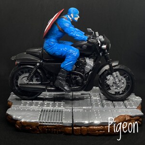 May include: A blue and red action figure of Captain America riding a black motorcycle with a silver base. The motorcycle has a silver exhaust pipe and a silver front wheel.