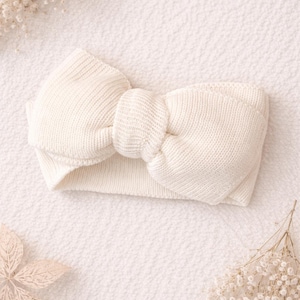 May include: Cream-coloured knitted headband featuring a large bow detail. The headband is made from a soft, textured material and is designed to be worn around the head. The bow is centrally placed, adding a decorative touch. A stylish accessory.