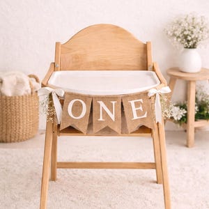 Puede incluir: Una trona de madera con una bandeja blanca y una pancarta que dice "ONE". La pancarta es de arpillera con letras blancas. La silla está decorada con flores blancas y cintas, ideal para una celebración de primer cumpleaños.