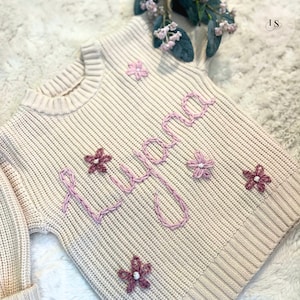 Puede incluir: Un suéter de punto color crema con el nombre "Lyana" bordado en hilo rosa. El suéter está decorado con flores bordadas en rosa y blanco.