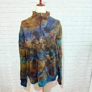 May include: A tie-dye pullover sweatshirt with a quarter-zip closure and a small red logo. The sweatshirt features a mix of colors, including brown, blue, and teal. The garment is displayed on a mannequin against a white brick wall.