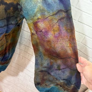May include: Close-up of a long-sleeved garment with a tie-dye pattern. The colors include shades of blue, purple, brown, and yellow. The fabric has a textured appearance. The sleeve is visible, with the cuff at the bottom.