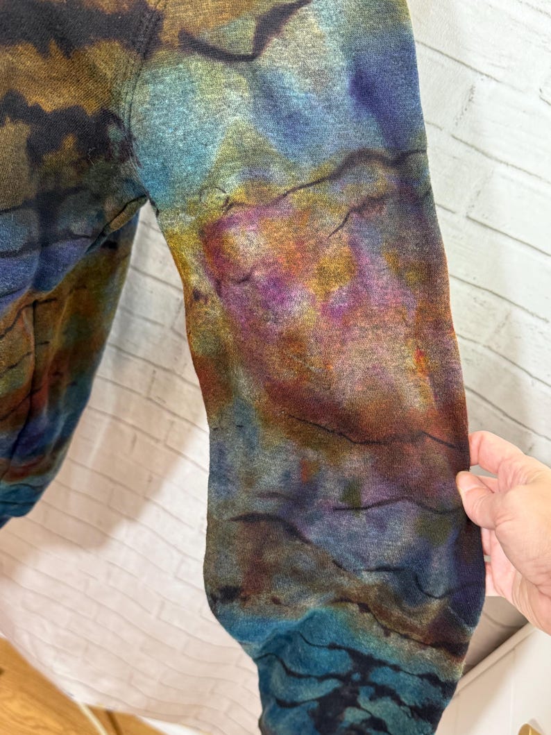 May include: Close-up of a long-sleeved garment with a tie-dye pattern. The colors include shades of blue, purple, brown, and yellow. The fabric has a textured appearance. The sleeve is visible, with the cuff at the bottom.