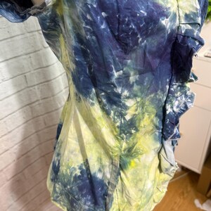 May include: A tie-dye shirt in shades of blue, yellow, and white. The shirt has short sleeves and a ruffled trim along the front. The fabric appears to be cotton or a similar lightweight material.