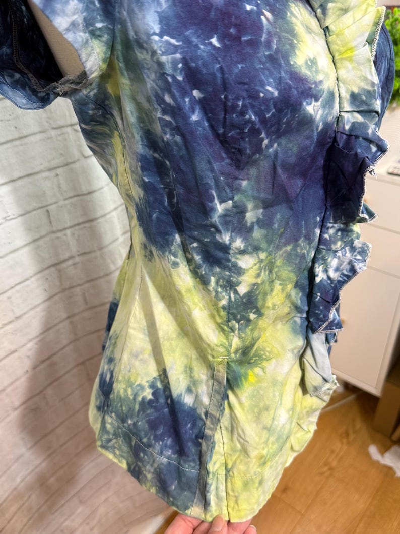 May include: A tie-dye shirt in shades of blue, yellow, and white. The shirt has short sleeves and a ruffled trim along the front. The fabric appears to be cotton or a similar lightweight material.