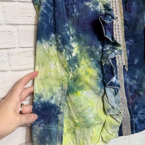 May include: A tie-dye jacket in shades of blue and yellow. The jacket features a ruffled trim and a decorative border. The fabric has a watercolor effect, with the colors blending together. The jacket is a unique piece of clothing.