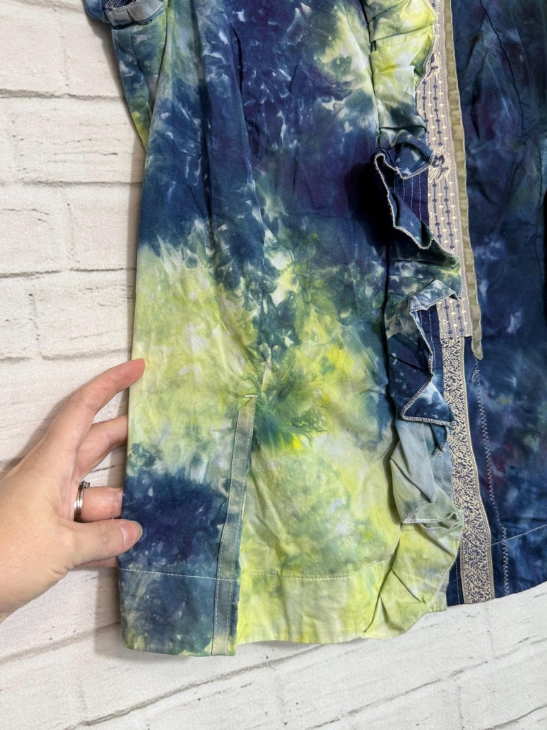 May include: A tie-dye jacket in shades of blue and yellow. The jacket features a ruffled trim and a decorative border. The fabric has a watercolor effect, with the colors blending together. The jacket is a unique piece of clothing.