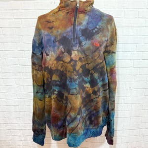 May include: A tie-dye pullover sweatshirt with a quarter-zip closure and a small logo on the chest. The colors include shades of blue, brown, and purple. The sweatshirt has long sleeves and a high collar. The garment is displayed on a mannequin.