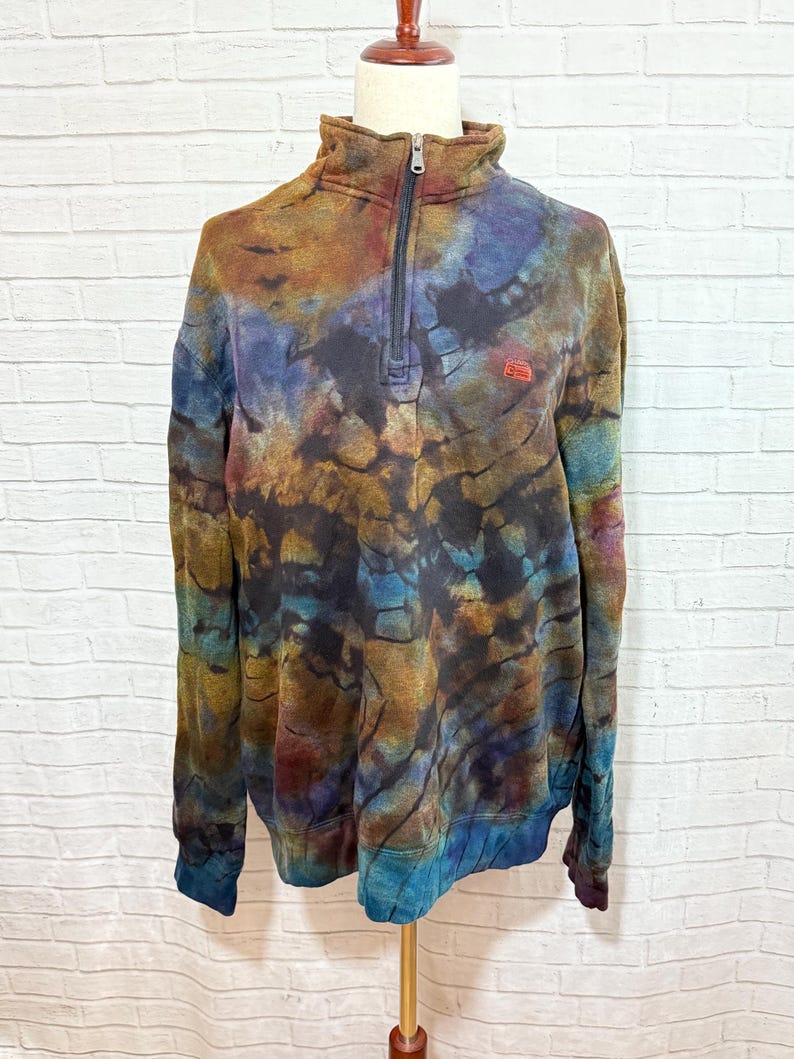 May include: A tie-dye pullover sweatshirt with a quarter-zip closure and a small logo on the chest. The colors include shades of blue, brown, and purple. The sweatshirt has long sleeves and a high collar. The garment is displayed on a mannequin.